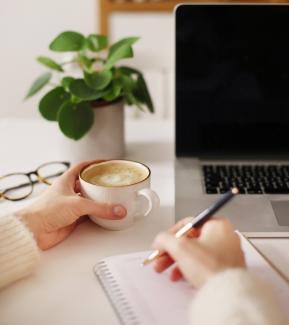 Point of view photo of a woman holding a pen on a notebook in her right hand, with a cappuccino in her left hand. A plant and a laptop in the background.