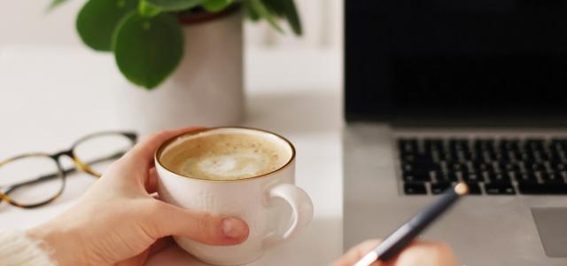 Point of view photo of a woman holding a pen on a notebook in her right hand, with a cappuccino in her left hand. A plant and a laptop in the background.