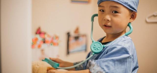 Image of a young child dressed up as a doctor, using a toy stethoscope on a stuffed animal.