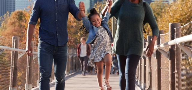 A young successful couple dressed casually and taking their daughter for a walk, lifting her up as they cross over a bridge.